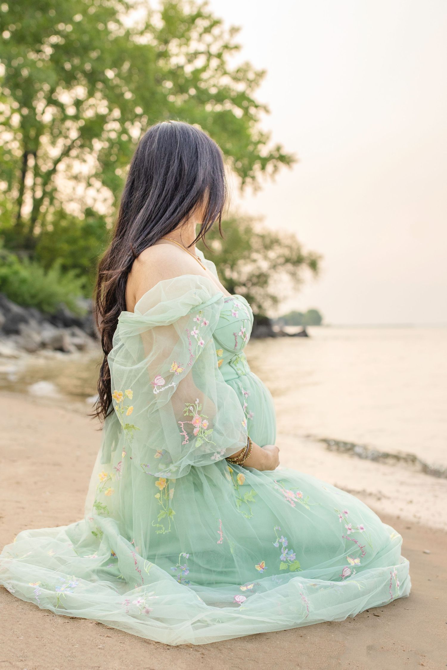 A person in a mint green floral dress sits contemplatively on a sandy beach at sunset.