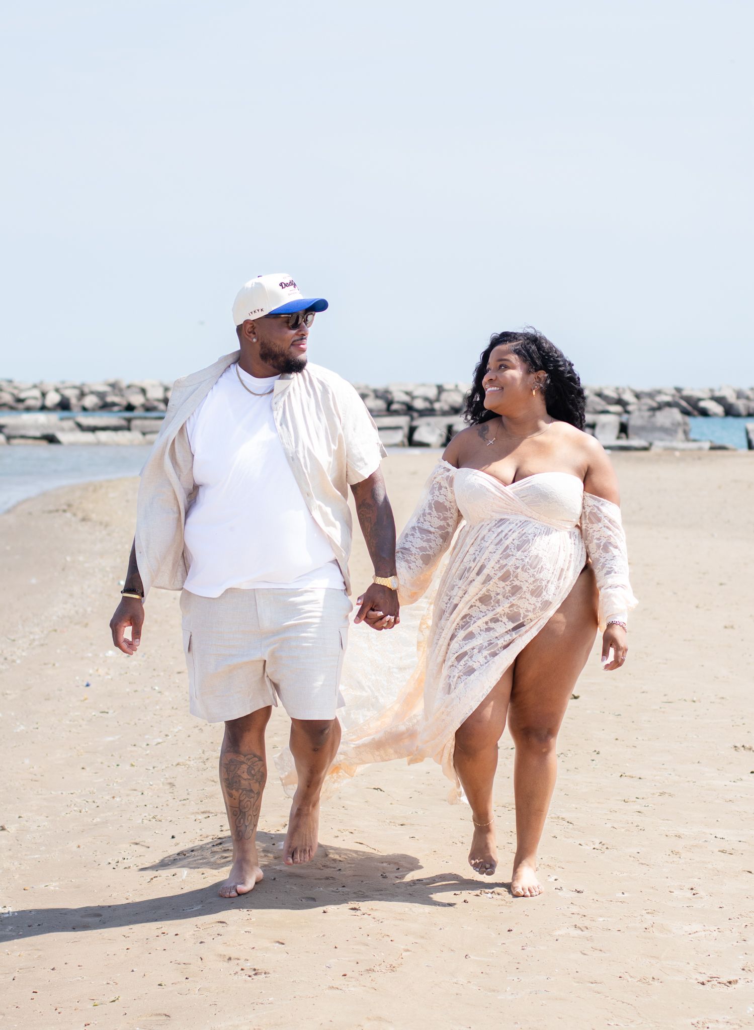 Couple walking hand in hand on beach wearing white outfits during sunny day.