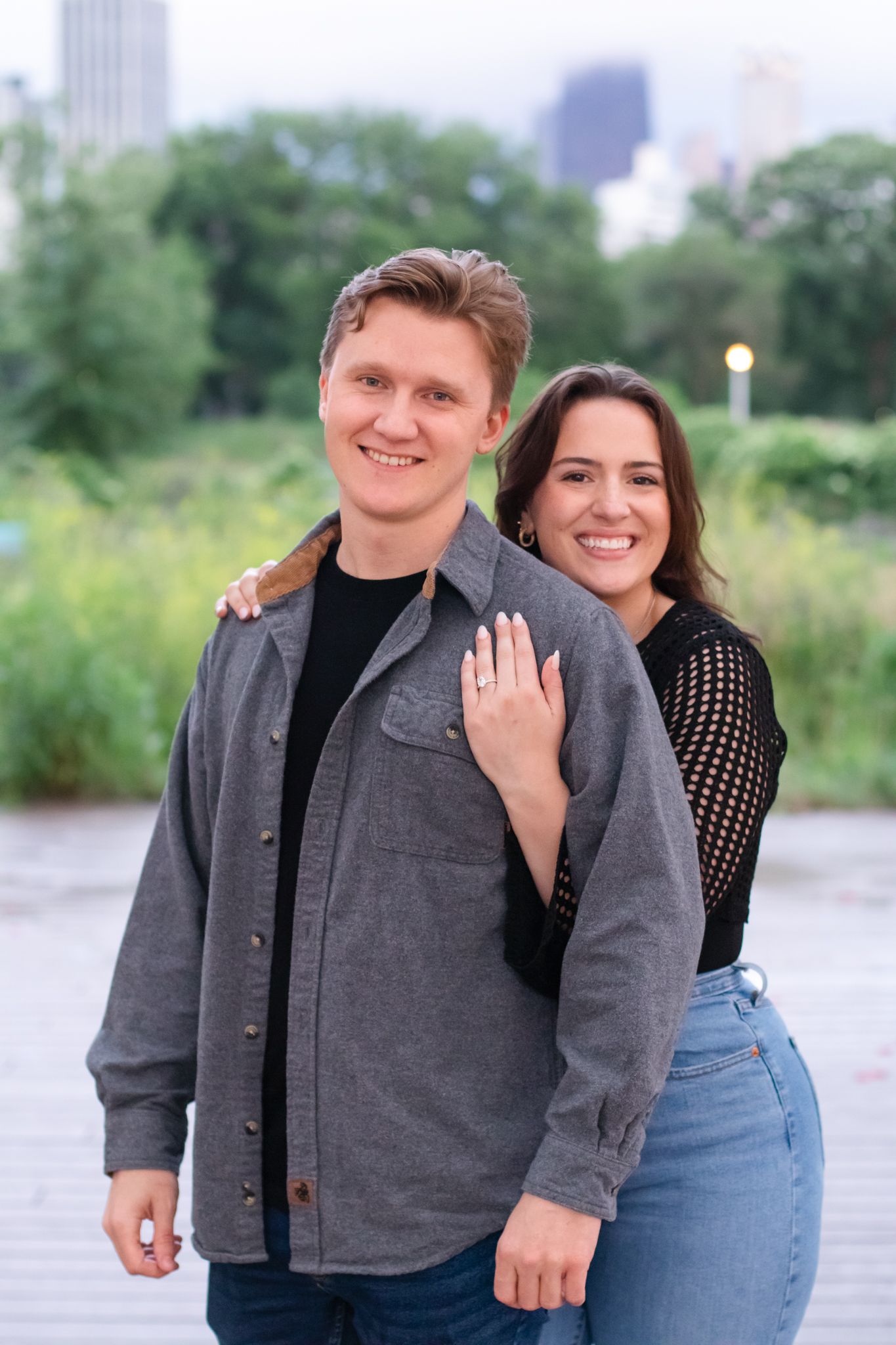 Happy couple poses for engagement photo with city skyline in background.
