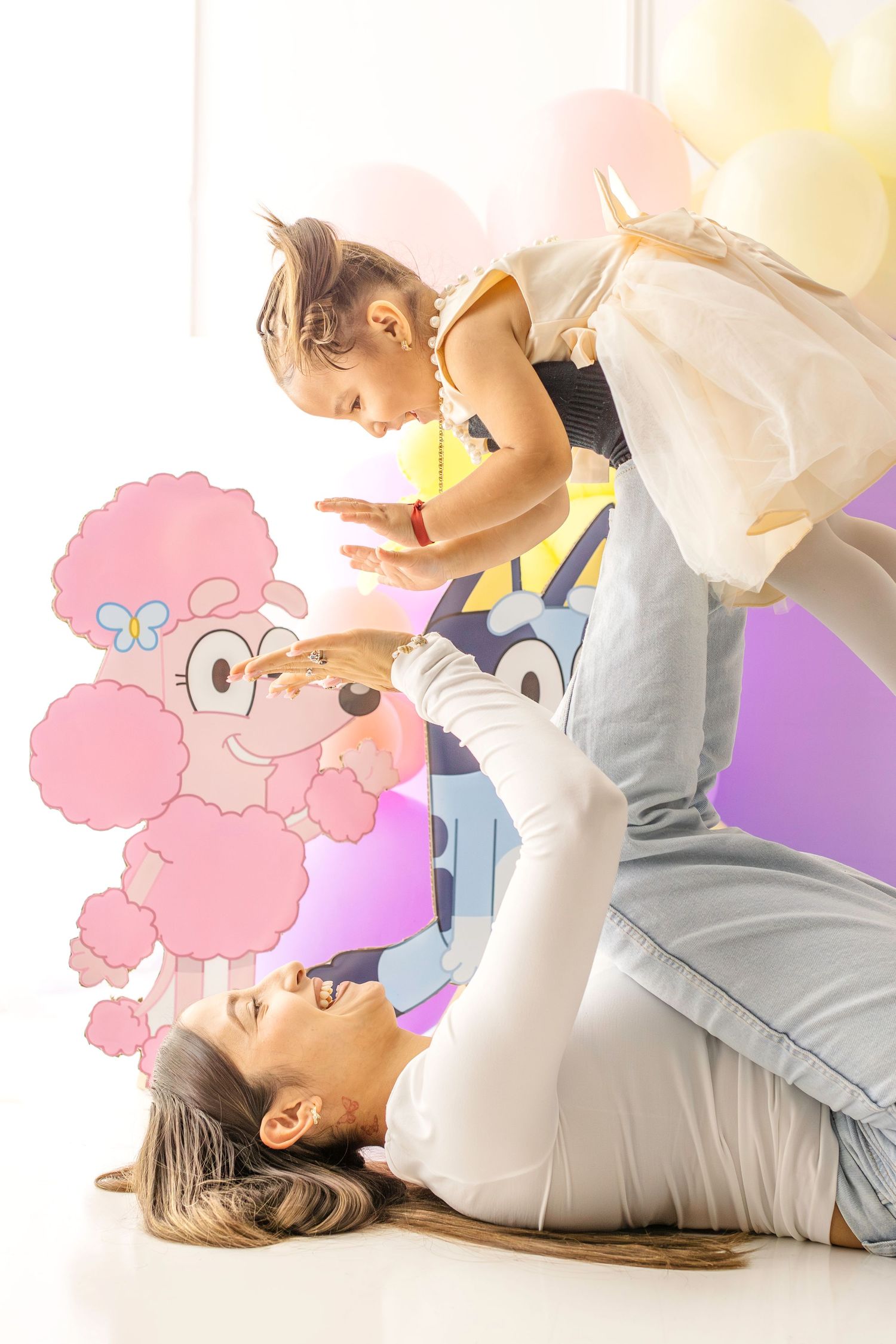 Mother lying on floor lifting toddler girl in the air, surrounded by colorful balloons and cartoon character illustrations.
