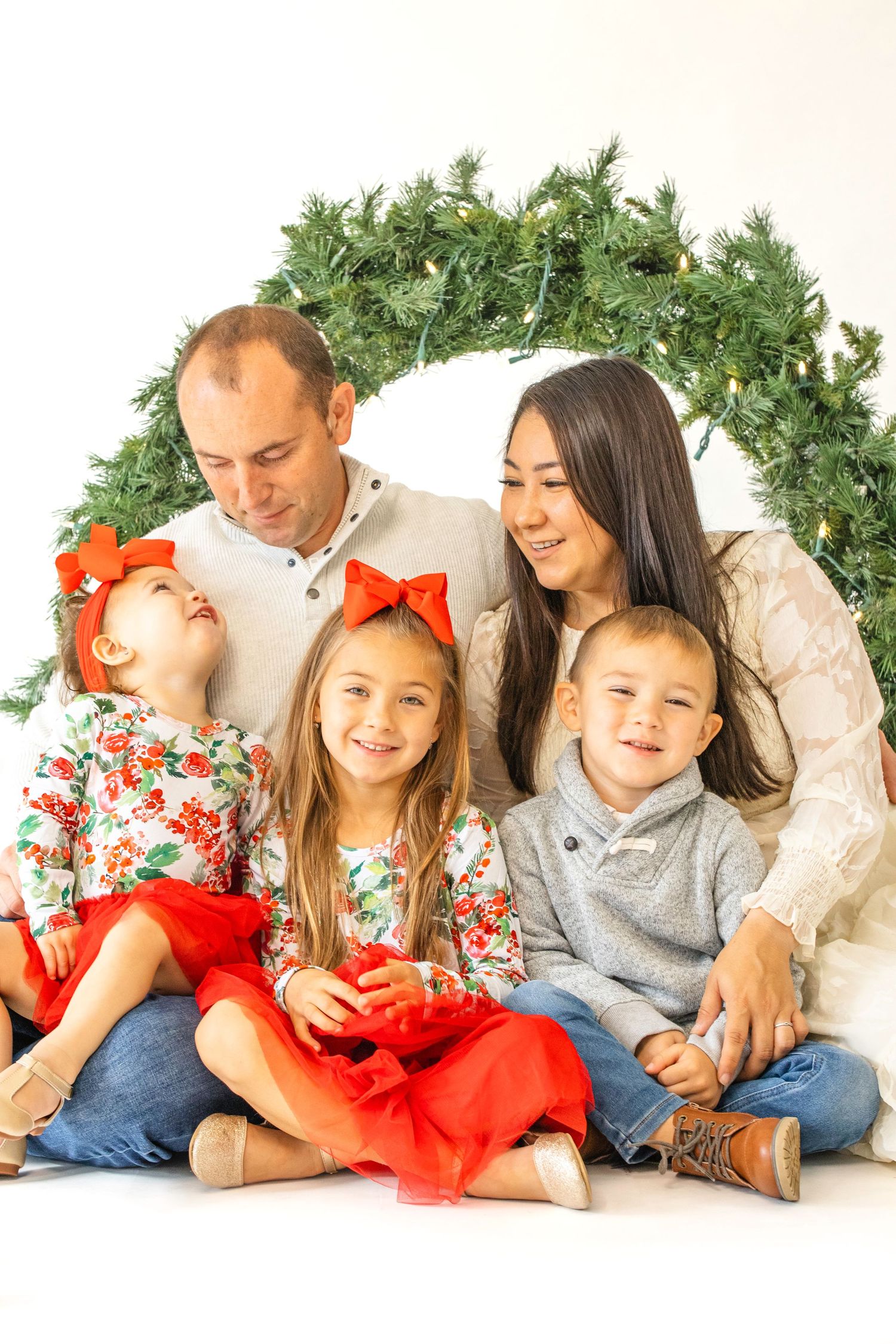 A family in coordinating white and red floral outfits lies together on a white backdrop with greenery decorations.
