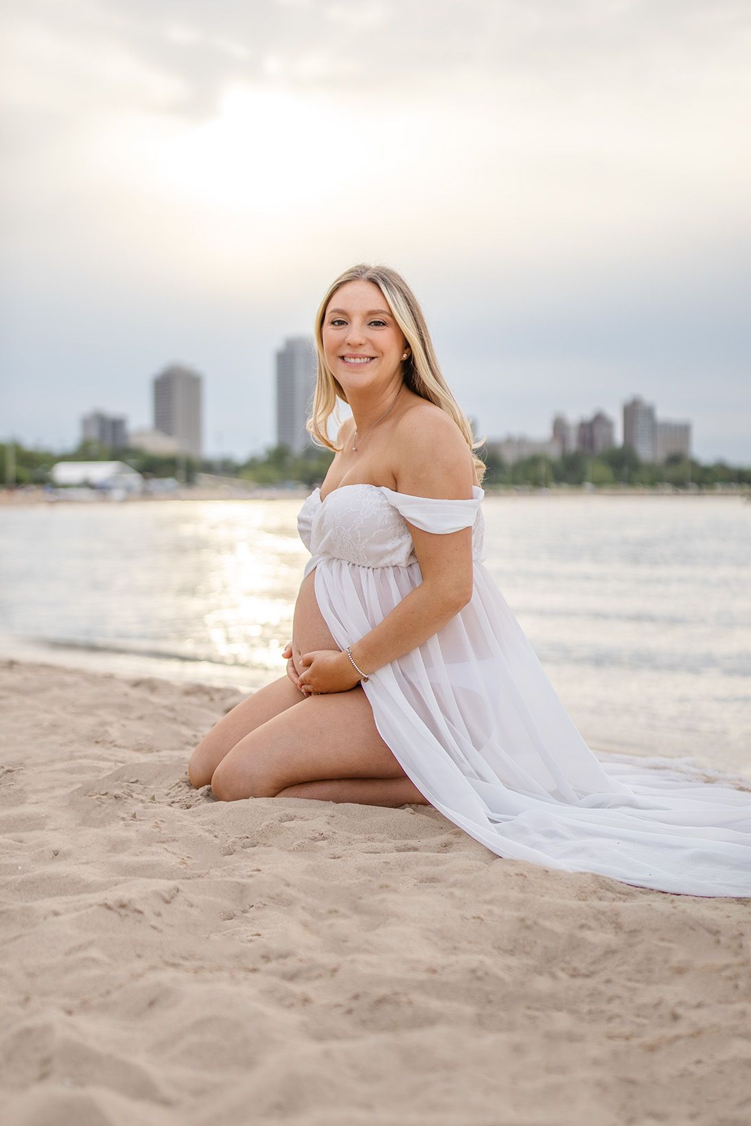 Pregnant woman in white off-shoulder gown sitting on sandy beach at sunset with city skyline in background.