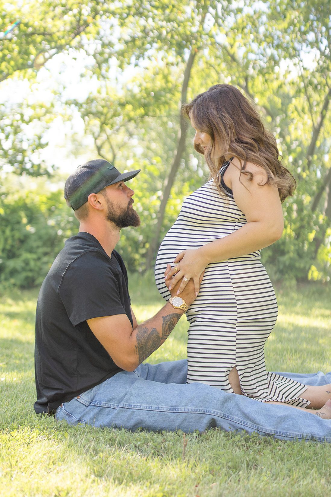 A man sits on grass touching a pregnant woman's belly during an outdoor maternity photoshoot.