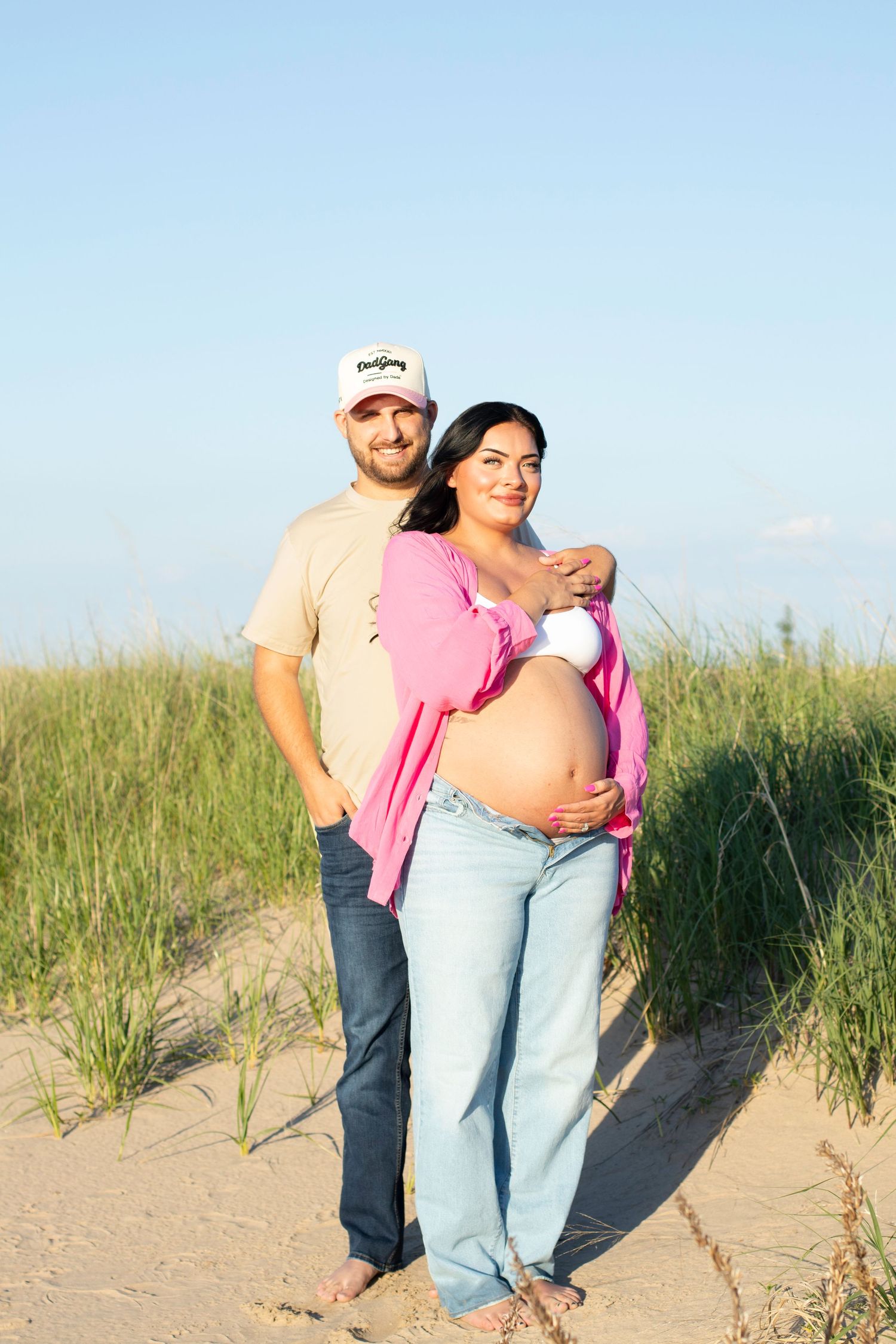Expecting couple poses together on sandy beach with pink cardigan.