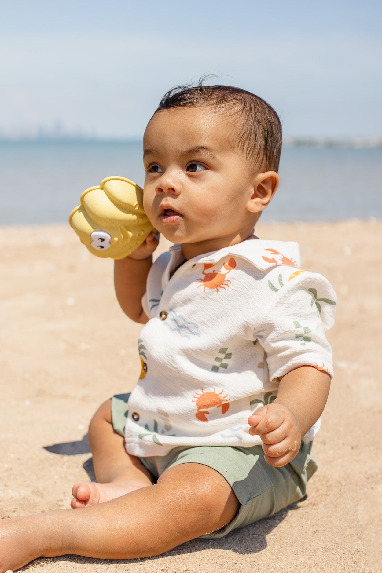 Cute baby sitting on sandy beach holding yellow toy, wearing white floral shirt and green shorts on sunny day.
