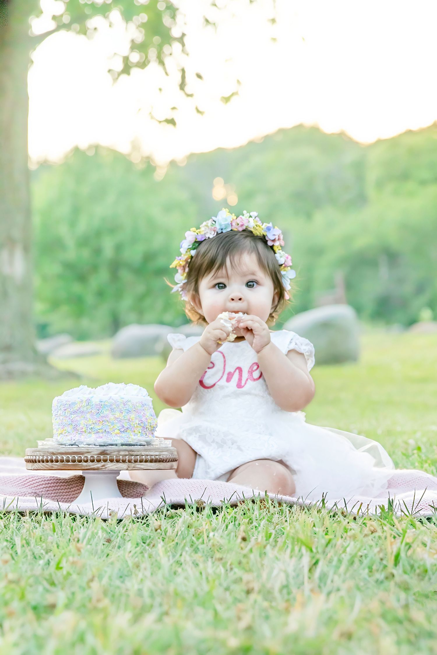 Sweet birthday celebration outdoors with smash cake on pink blanket and floral crown against dreamy natural backdrop.