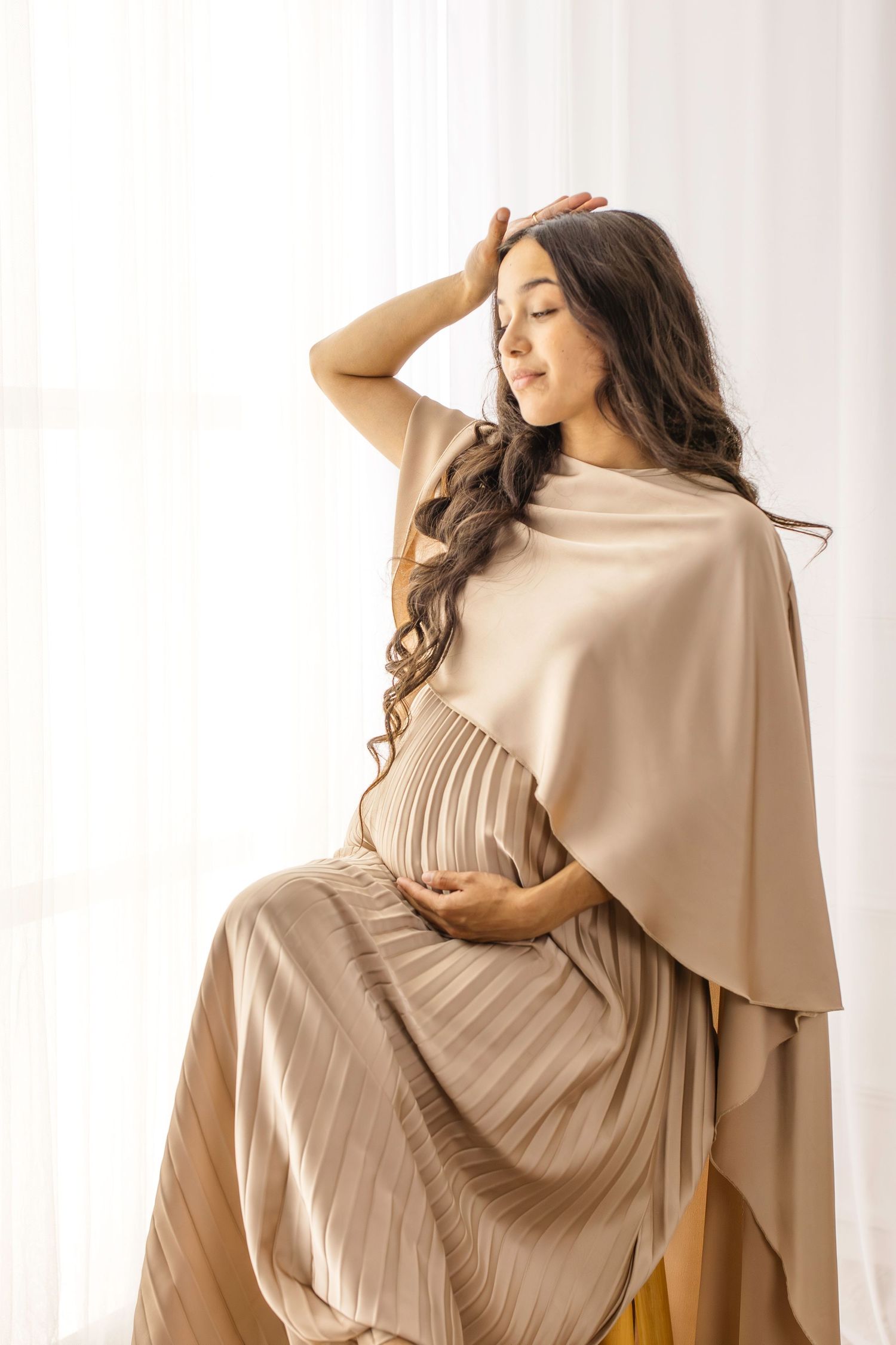 Model wearing a beige knit dress with asymmetrical cape overlay poses elegantly against white curtain backdrop.