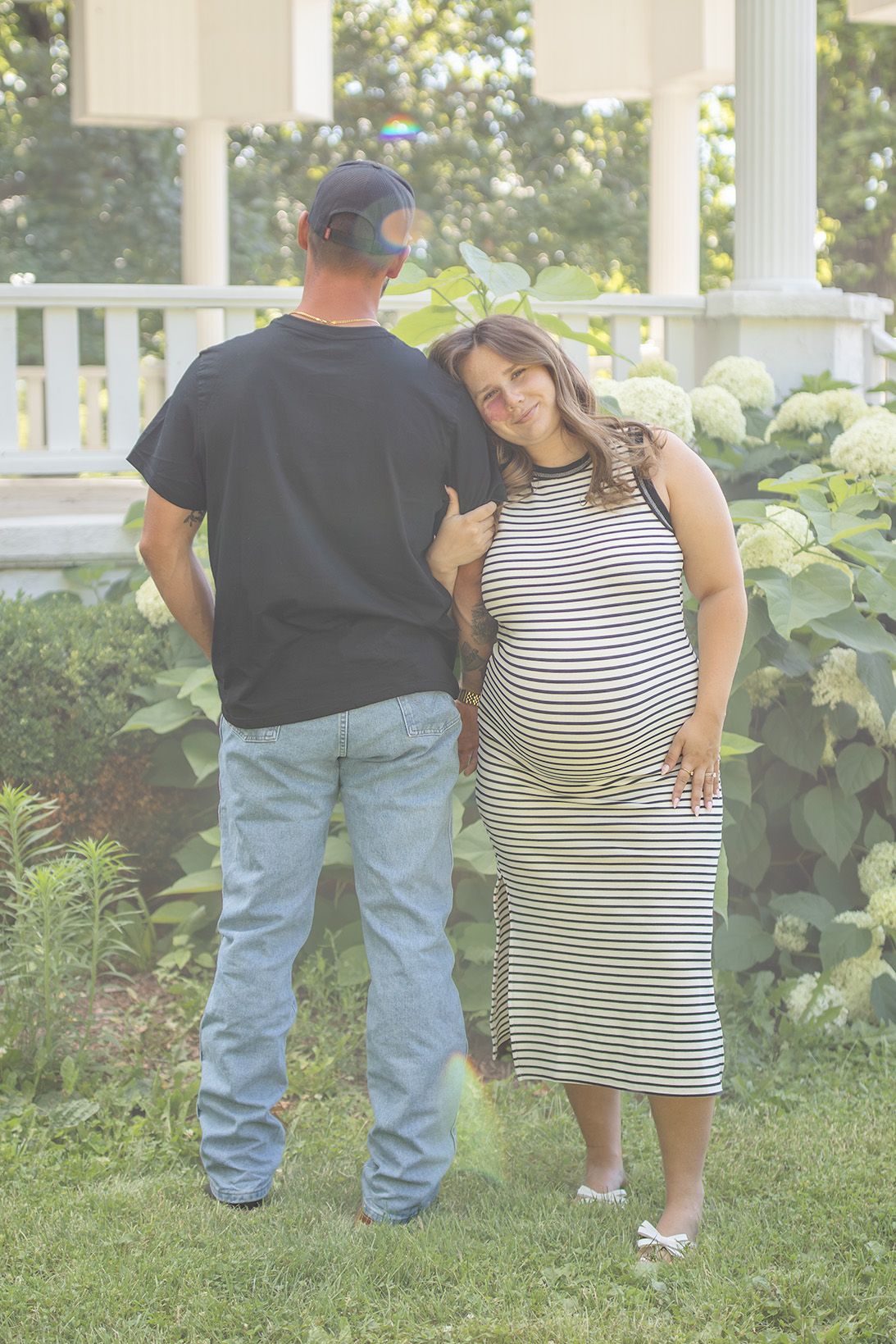 Pregnant woman in striped dress leaning on mans shoulder, standing in garden near white hydrangeas.