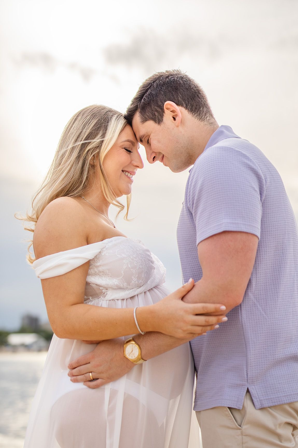 Couple sharing a tender forehead touch on the beach, woman in white off-shoulder dress, man in light blue shirt.