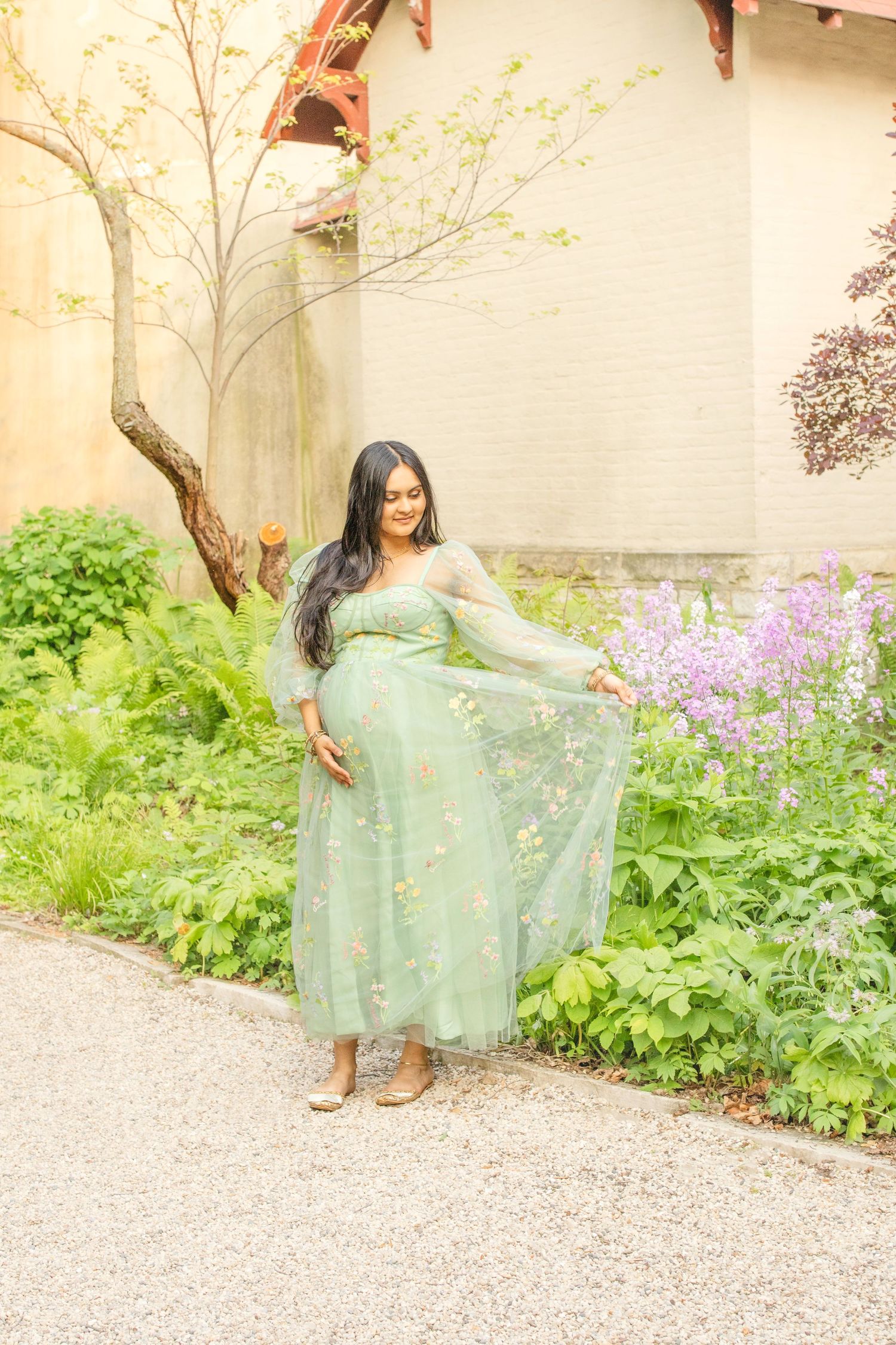 A person poses gracefully in a mint green tulle dress surrounded by blooming flowers and trees in a garden setting.