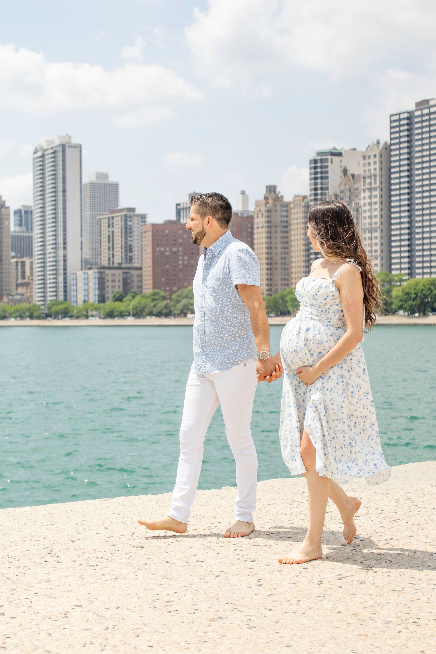 A couple walks hand in hand along the beach near Lake Michigan with the Chicago skyline rising in the background.