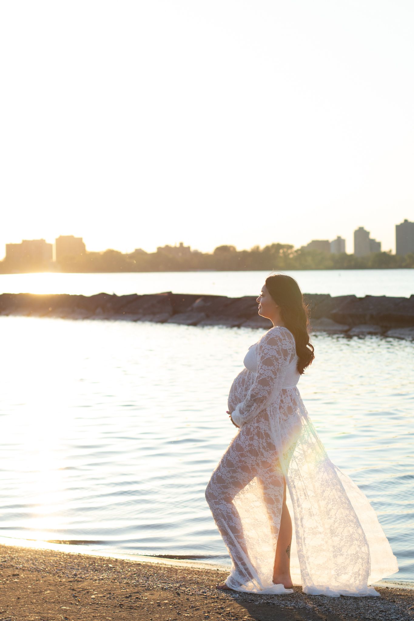 Silhouette of pregnant person in lace dress against sunset waterfront.