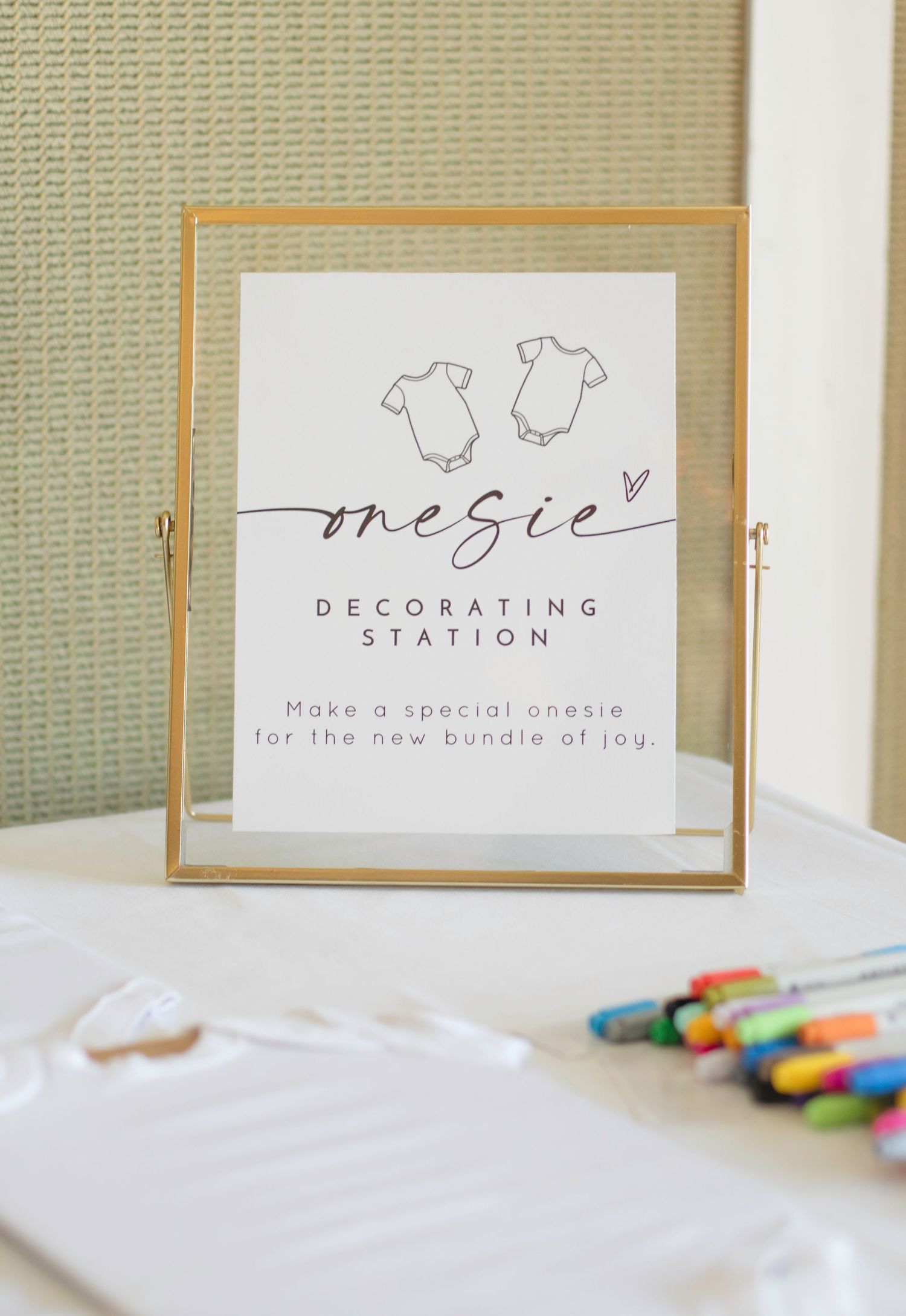 A decorating station sign with baby onesie illustrations and elegant script text in a gold frame.