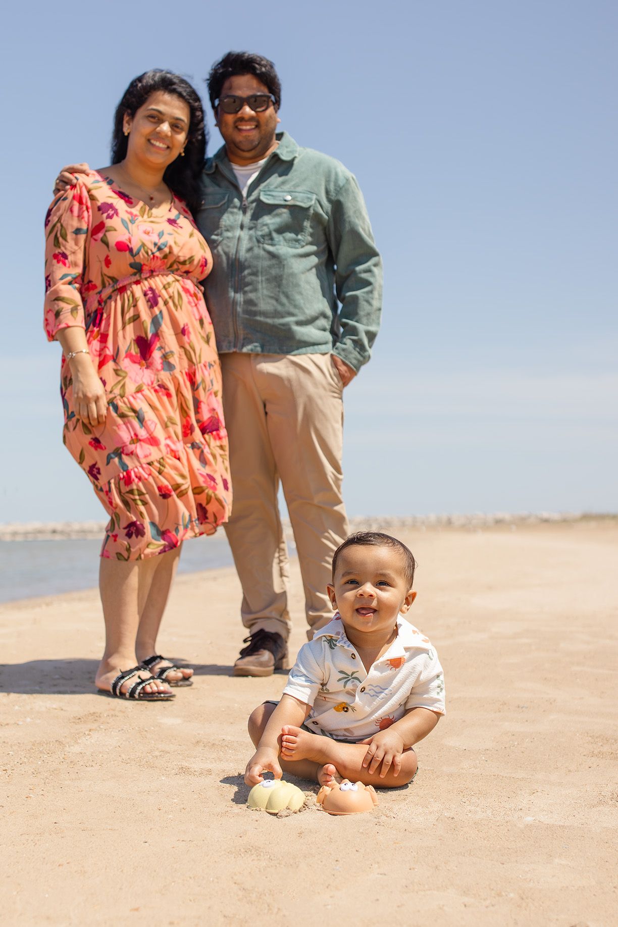 Happy family with baby on sunny beach, parents smiling behind toddler playing in sand near water.