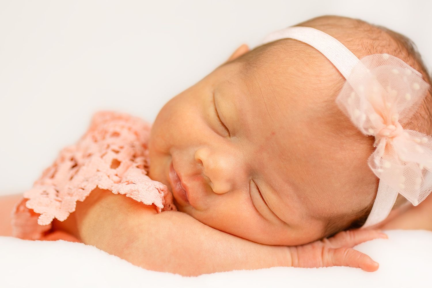 Sleeping newborn baby girl in pink crochet outfit and white bow headband on white background.