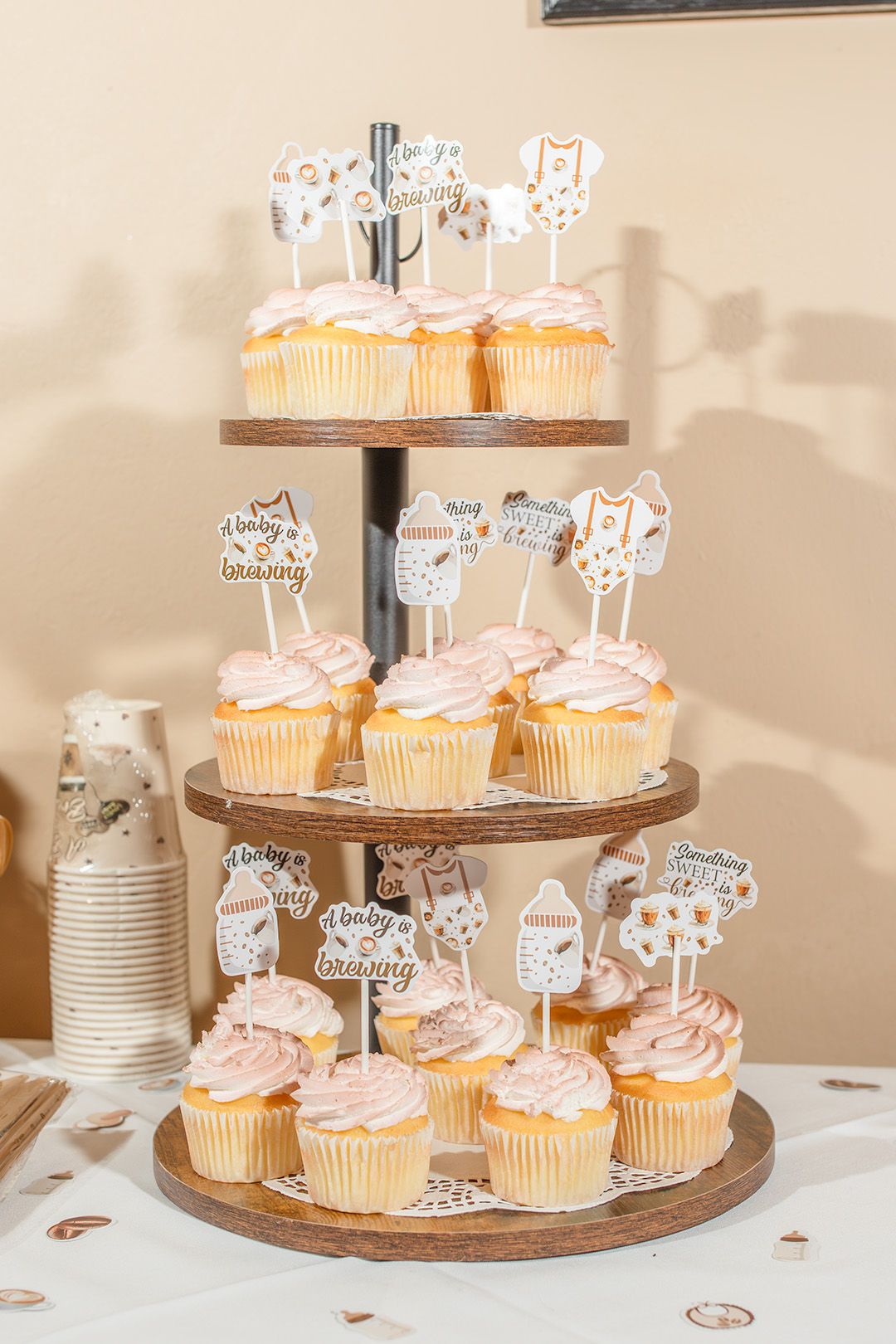 Three-tier wooden cupcake stand with pink frosted cupcakes topped with baby shower decorations.