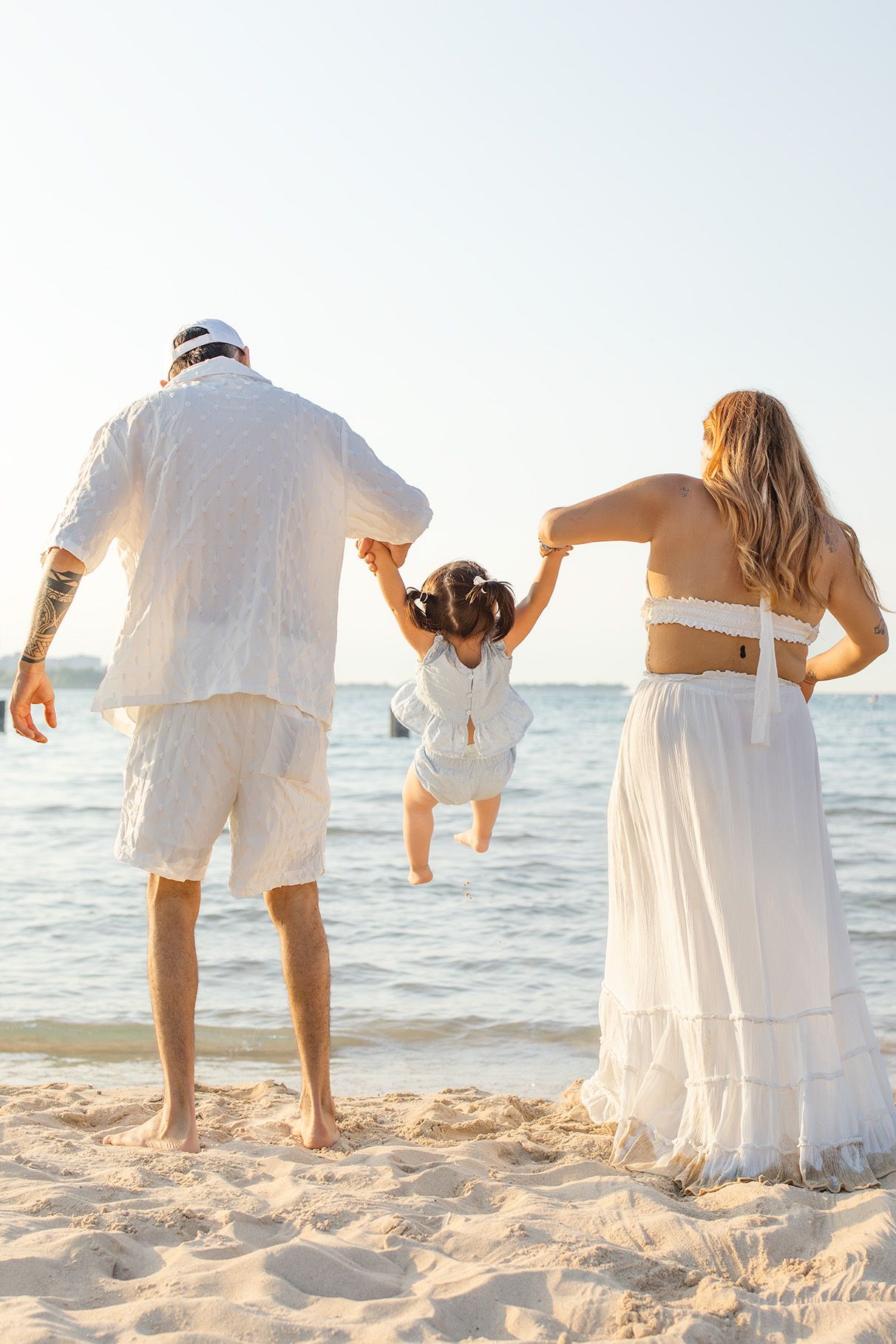 Family in white outfits swinging their child on a sunny beach.