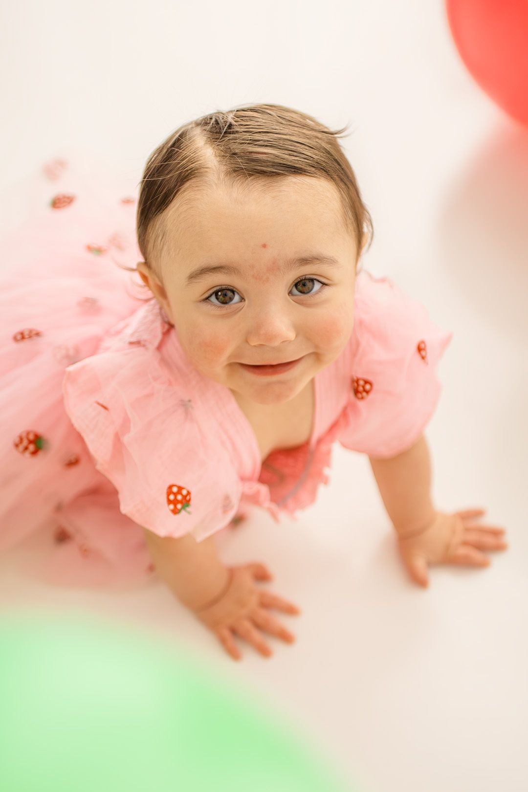 A smiling baby girl in a pink tutu dress with strawberry details crawls on a white floor surrounded by colorful balloons.