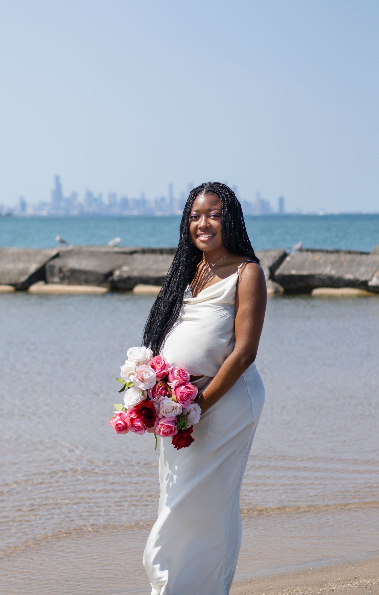 Expecting mother in white dress holds pink roses during beach photoshoot with city backdrop.