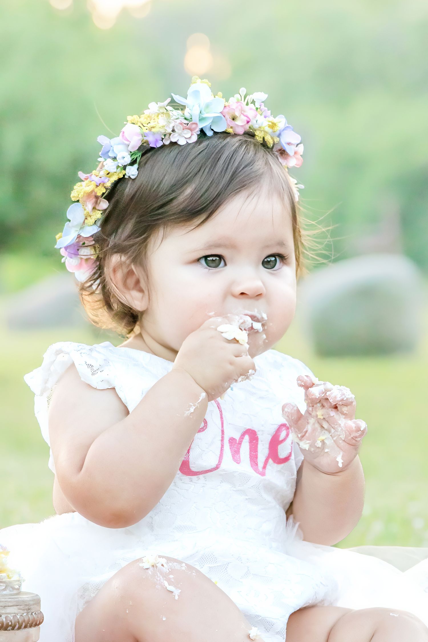 Baby girl in floral crown eating cake smash on her first birthday outdoors in white dress.