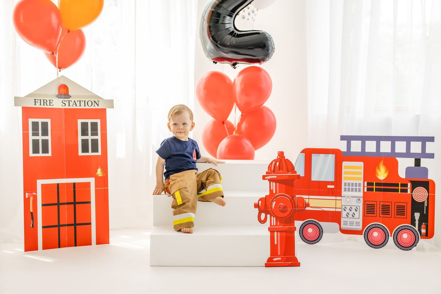Toddler in firefighter boots sits on white steps surrounded by fire station decorations and red balloons at a birthday party.