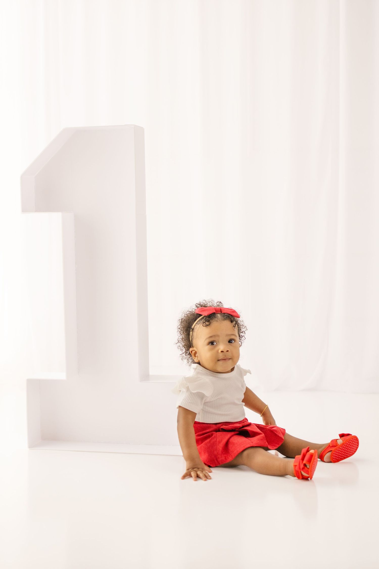 Cute baby girl in red skirt and bow headband sits beside a large number one prop for first birthday photos.