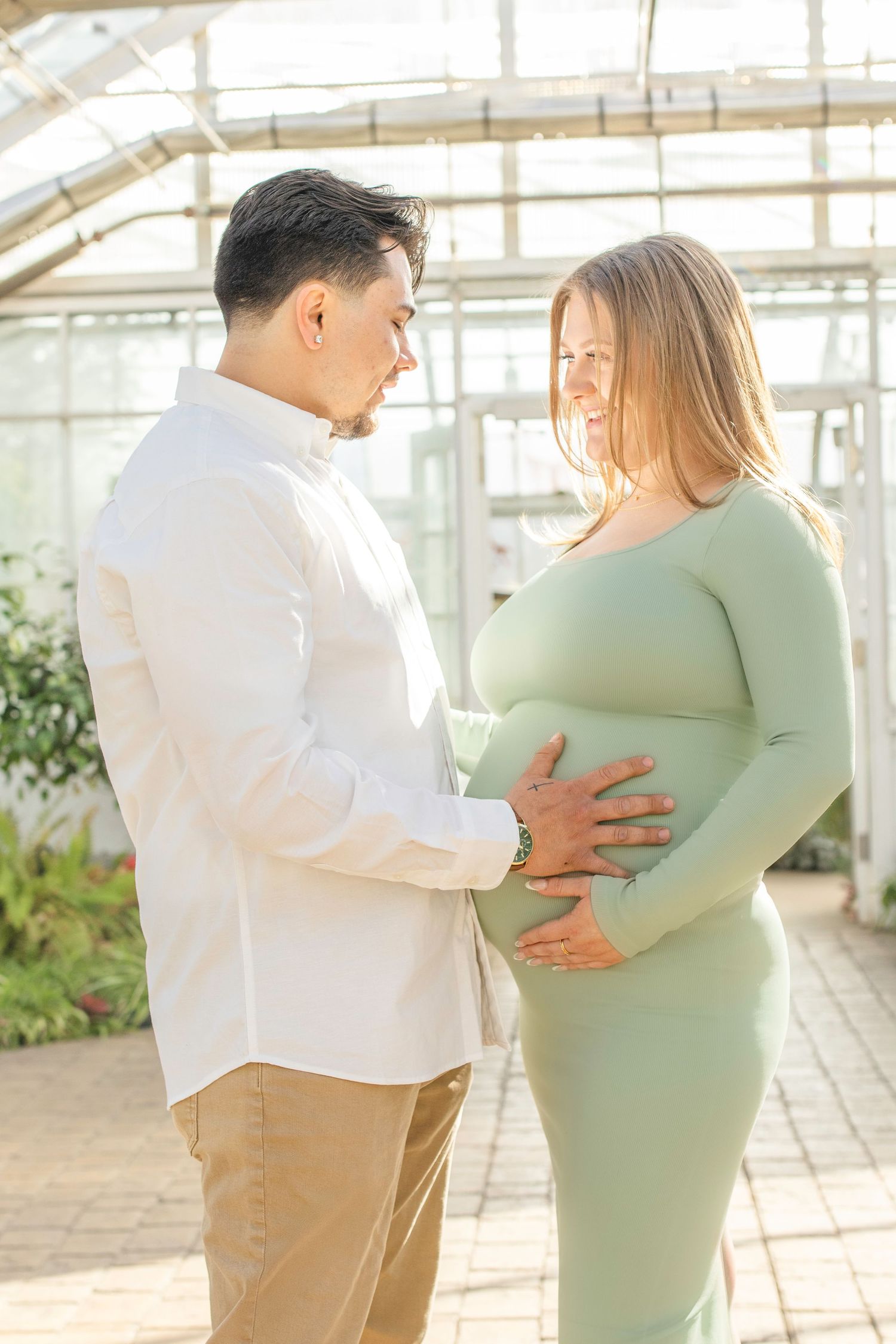 Intimate maternity photo of couple in white and sage green attire inside a sunlit greenhouse.