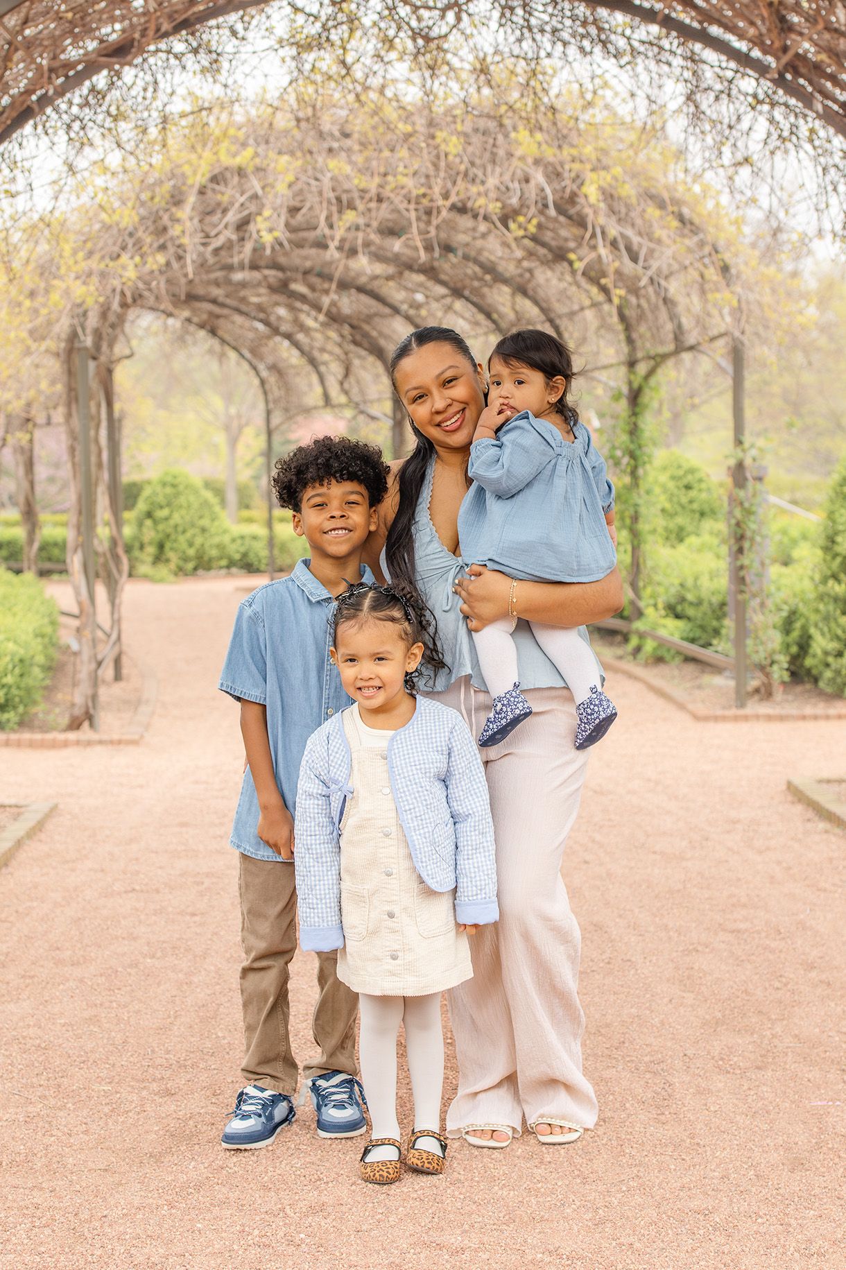 A mother smiles holding her toddler while posing with her two children under a garden archway.