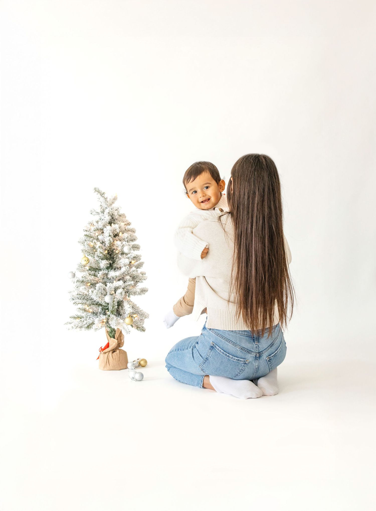 A person in a white sweater and jeans sits beside a small decorated Christmas tree on a white backdrop.