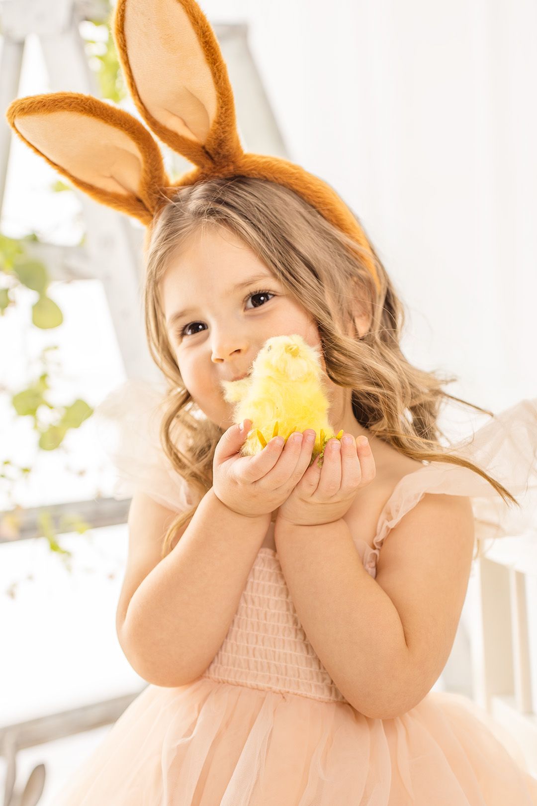 Young child in pink dress and bunny ears plays with yellow chick toy in sunny setting.