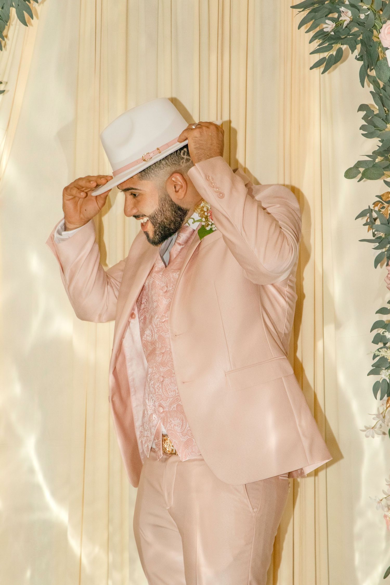 Person in a light pink formal suit poses playfully against a beige curtain backdrop.