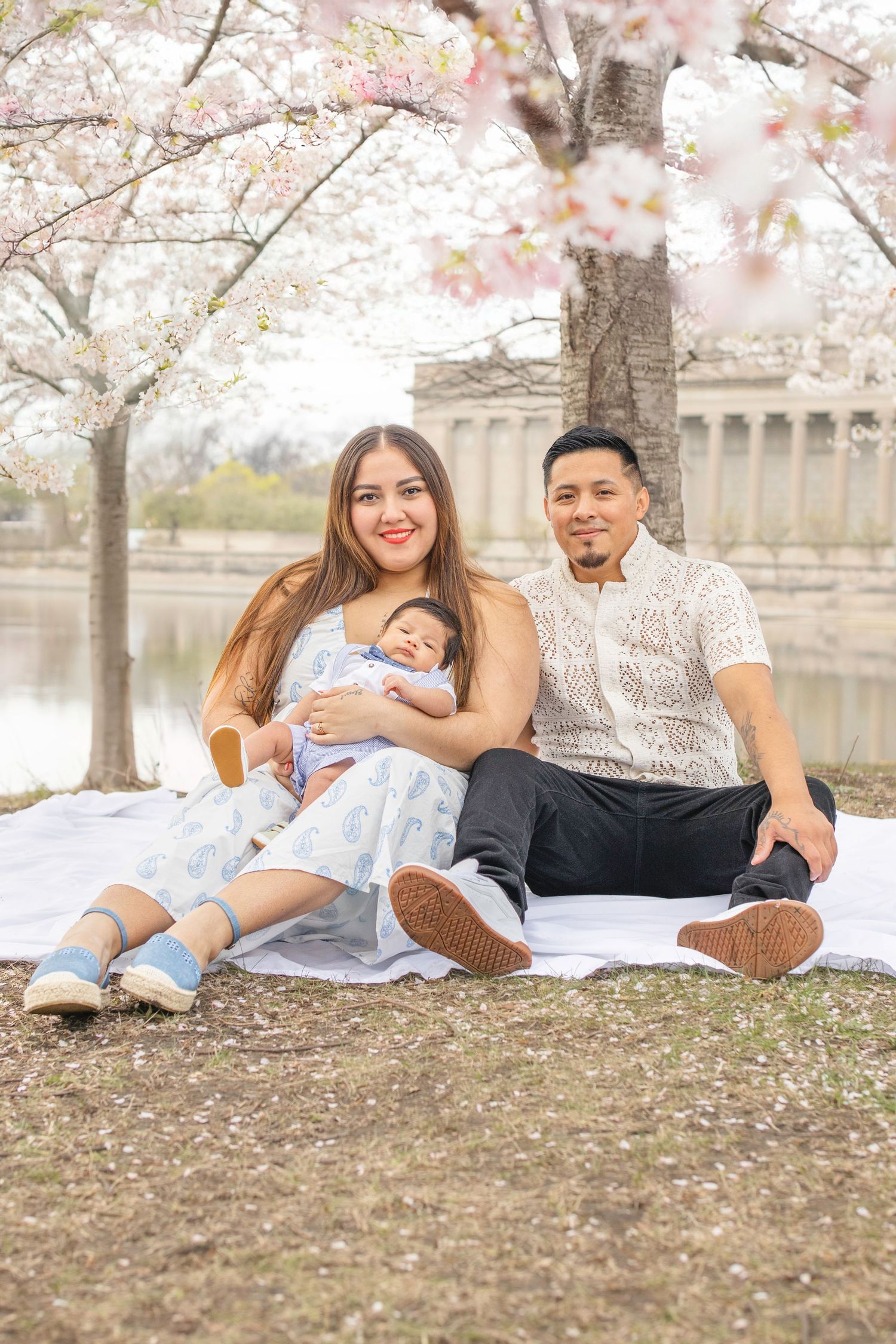 Family sits on picnic blanket under blooming cherry blossom trees near a reflective pond on peaceful spring day.