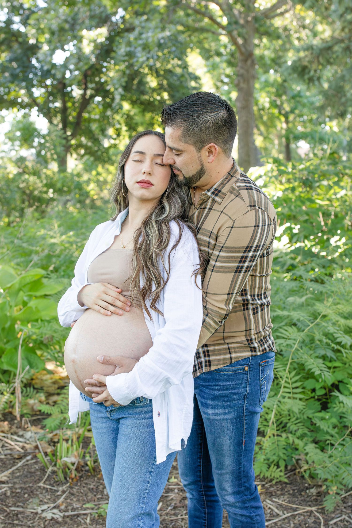 A tender moment captured outdoors as an expecting couple embraces in a sunlit woodland setting.