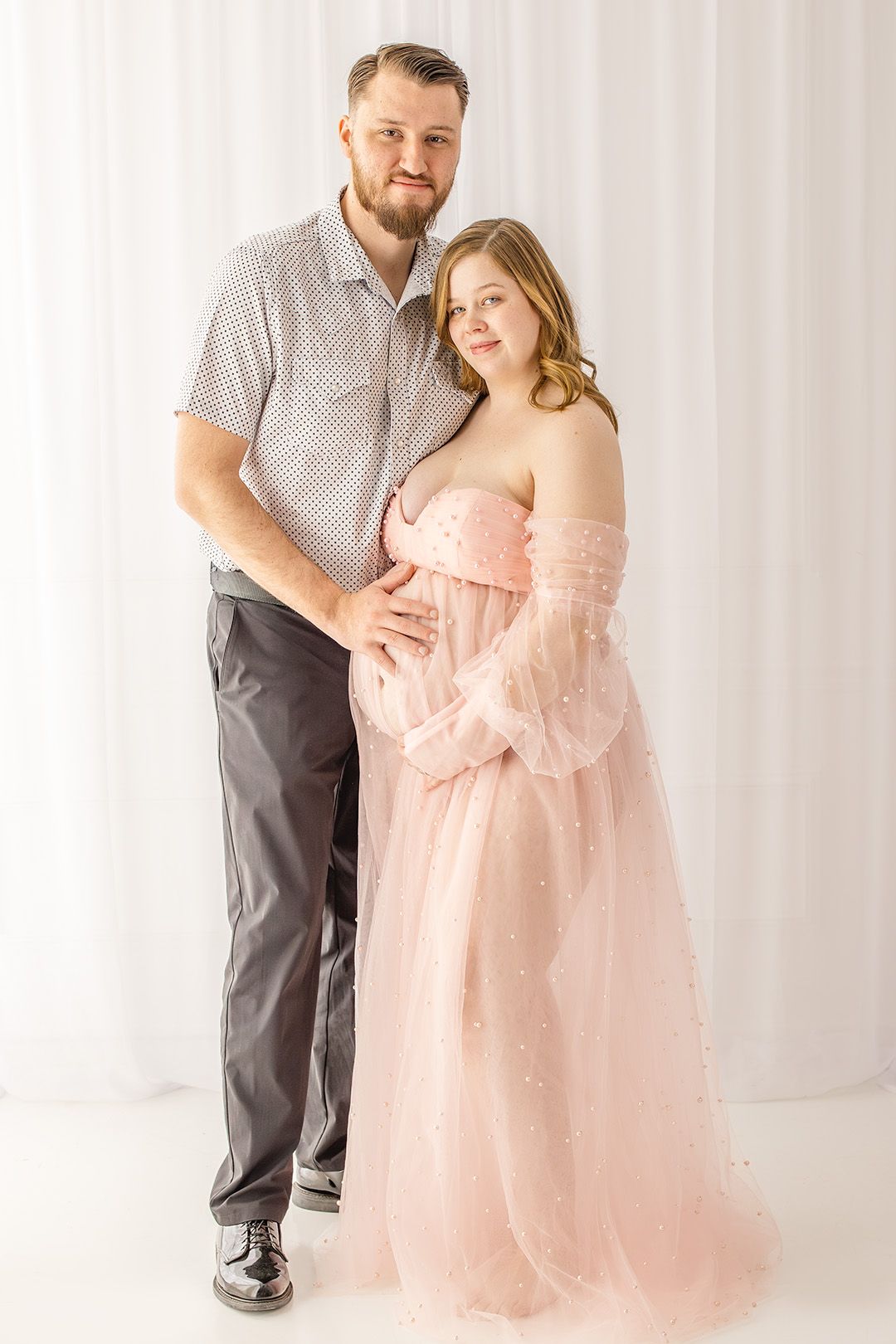 A couple in a romantic maternity photo shoot wearing pink and grey against a white backdrop.