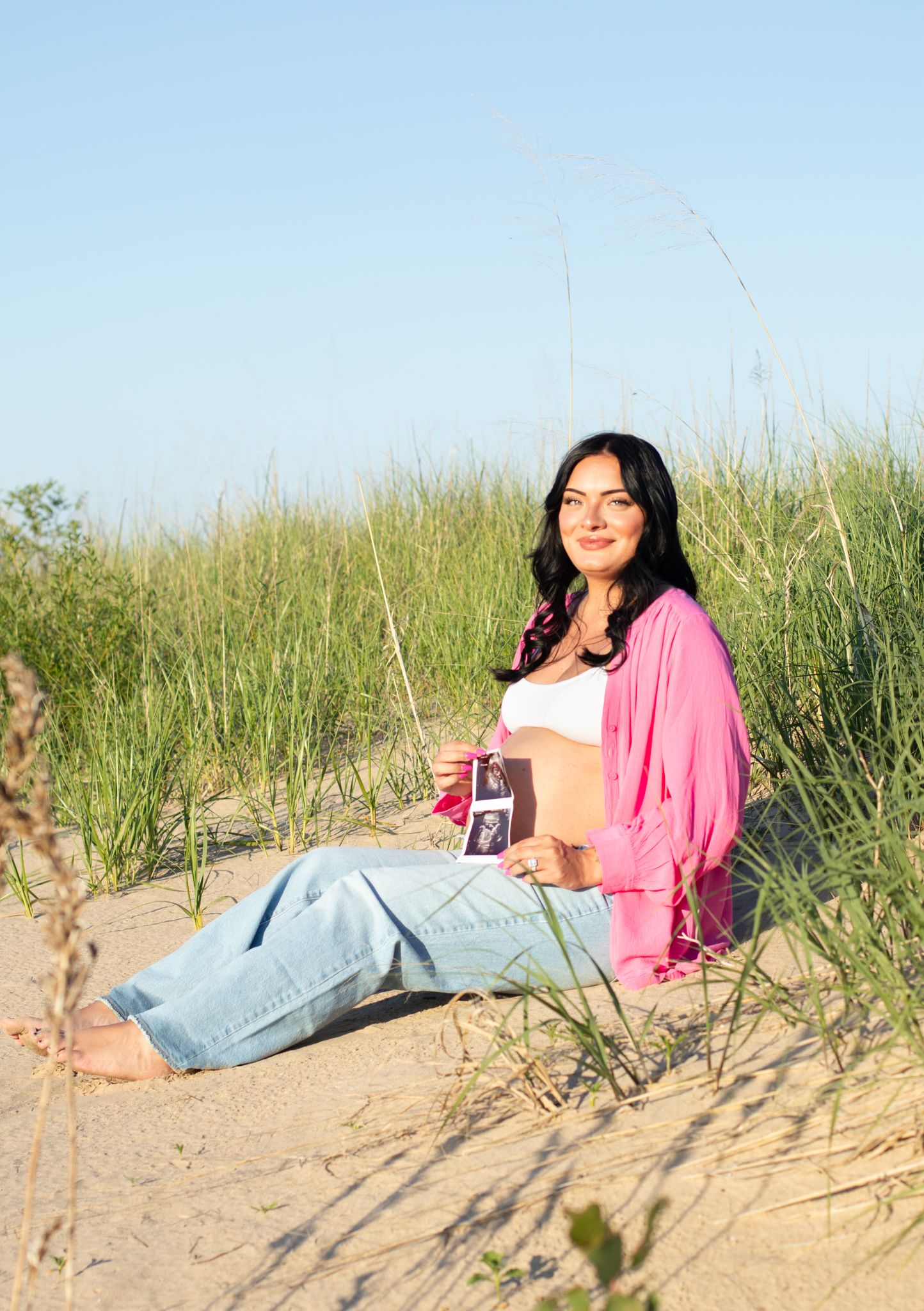Maternity photo session on sandy beach with pink cardigan and white top.