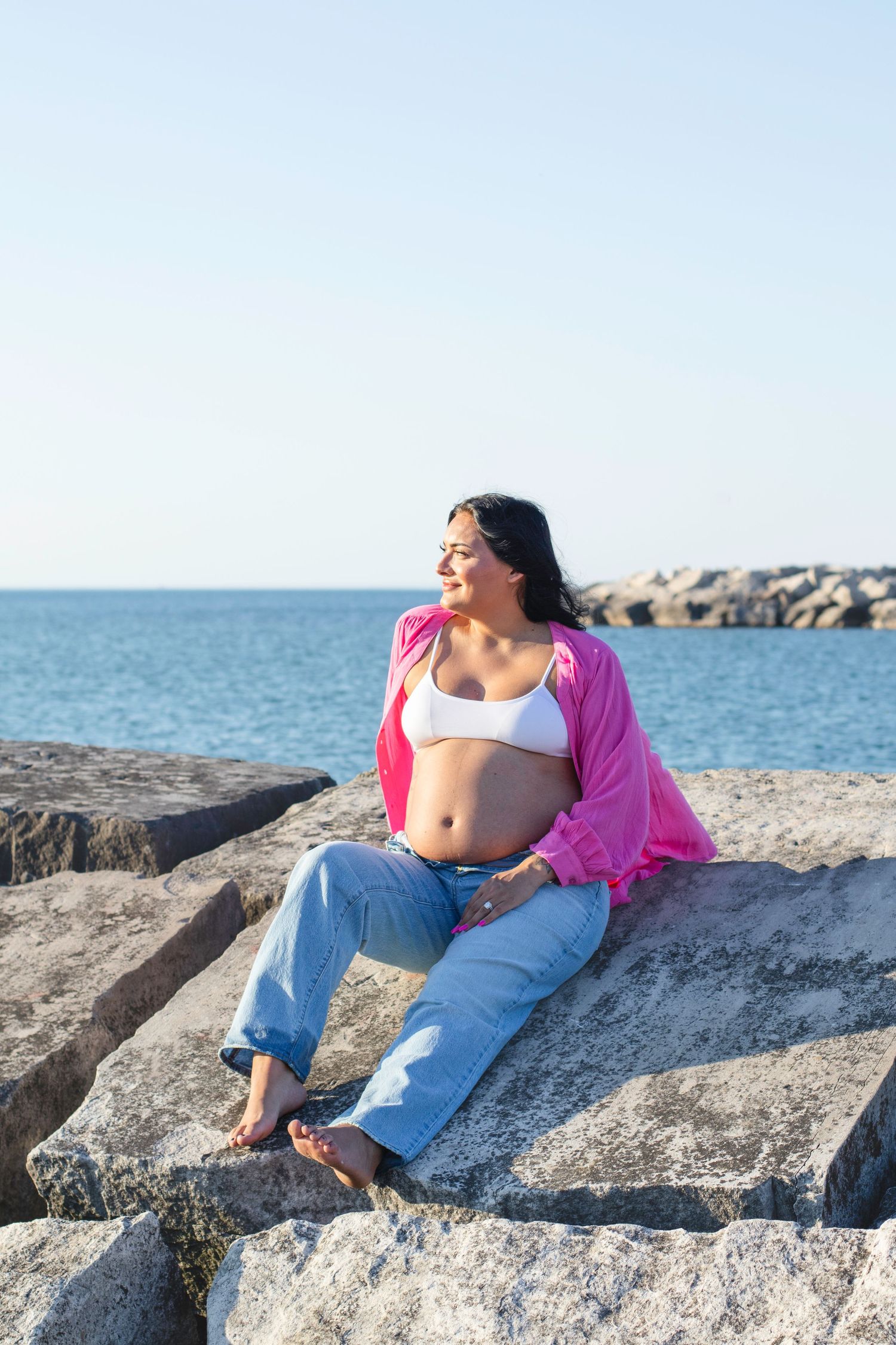 Person in pink cardigan and jeans relaxing on rocky beach overlook near ocean.