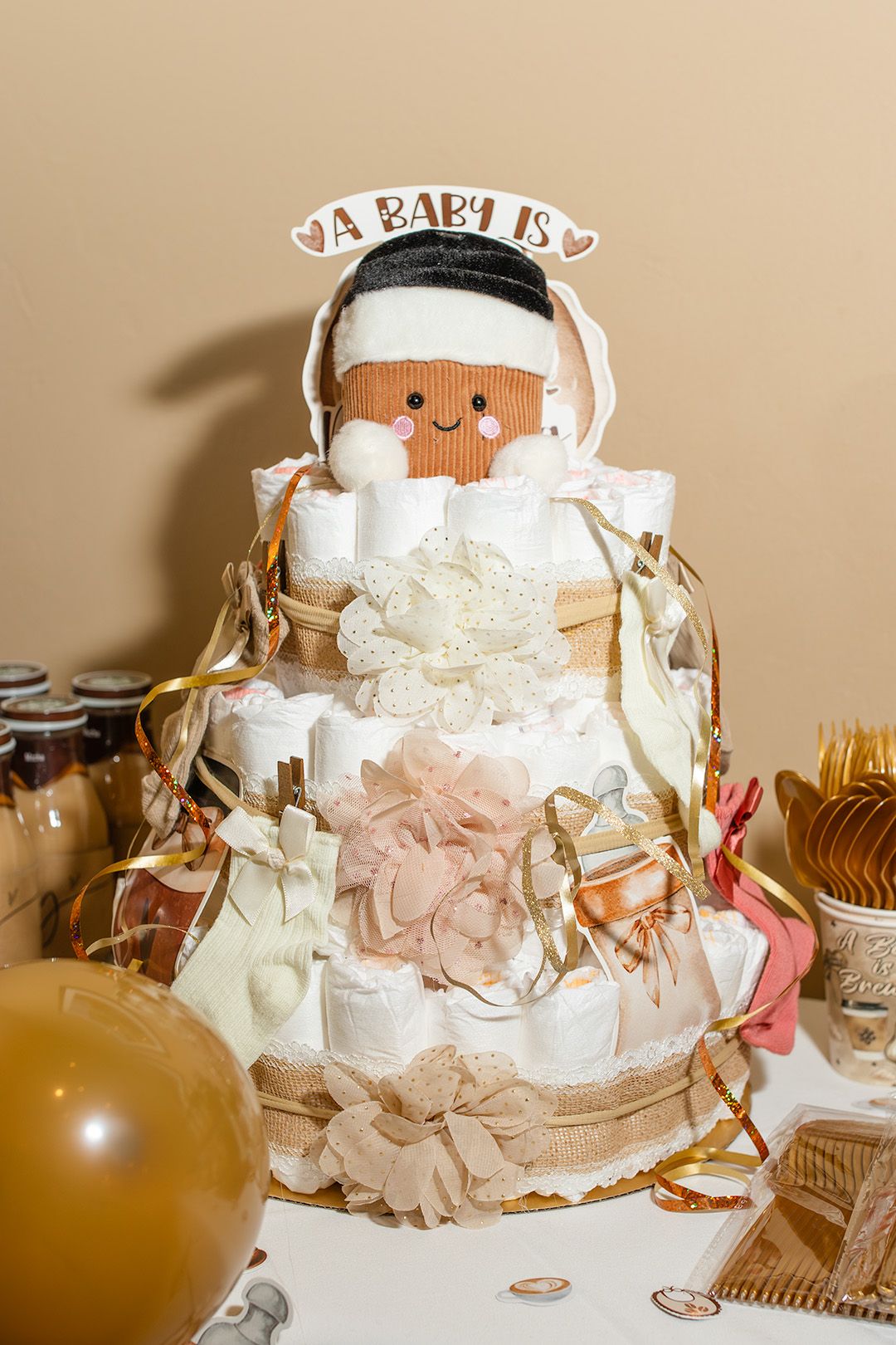 A decorative diaper cake topped with a baby doll figurine and an A Baby Is sign, adorned with gold ribbons and bows.