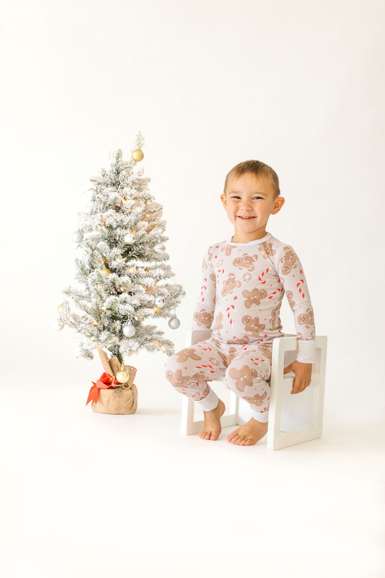 A small child in pink floral pajamas sits on white blocks next to a frosted Christmas tree in a bright studio setting.