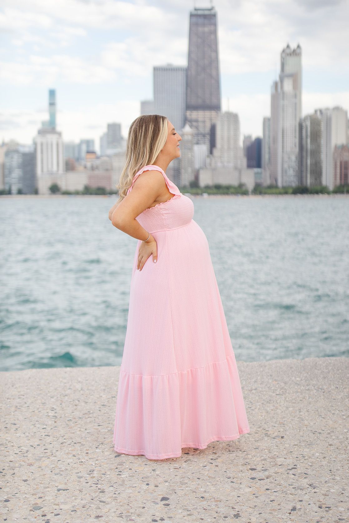 Pregnant woman in pink maxi dress poses by Chicago lakefront with city skyline backdrop.