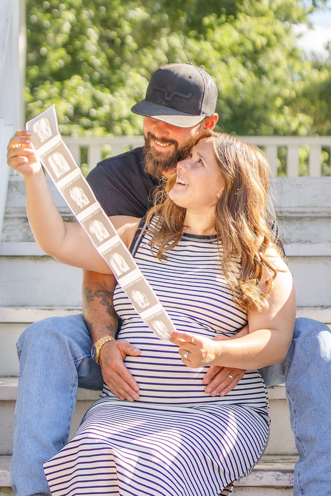 Pregnant couple sitting on steps holding ultrasound photos, man in cap embracing woman in striped dress outdoors.