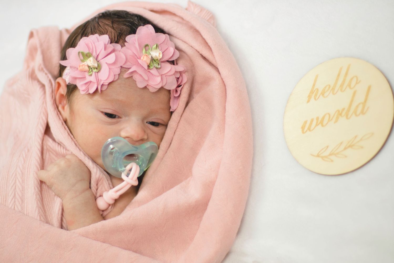 Newborn with pacifier wrapped in pink blanket next to hello world sign.