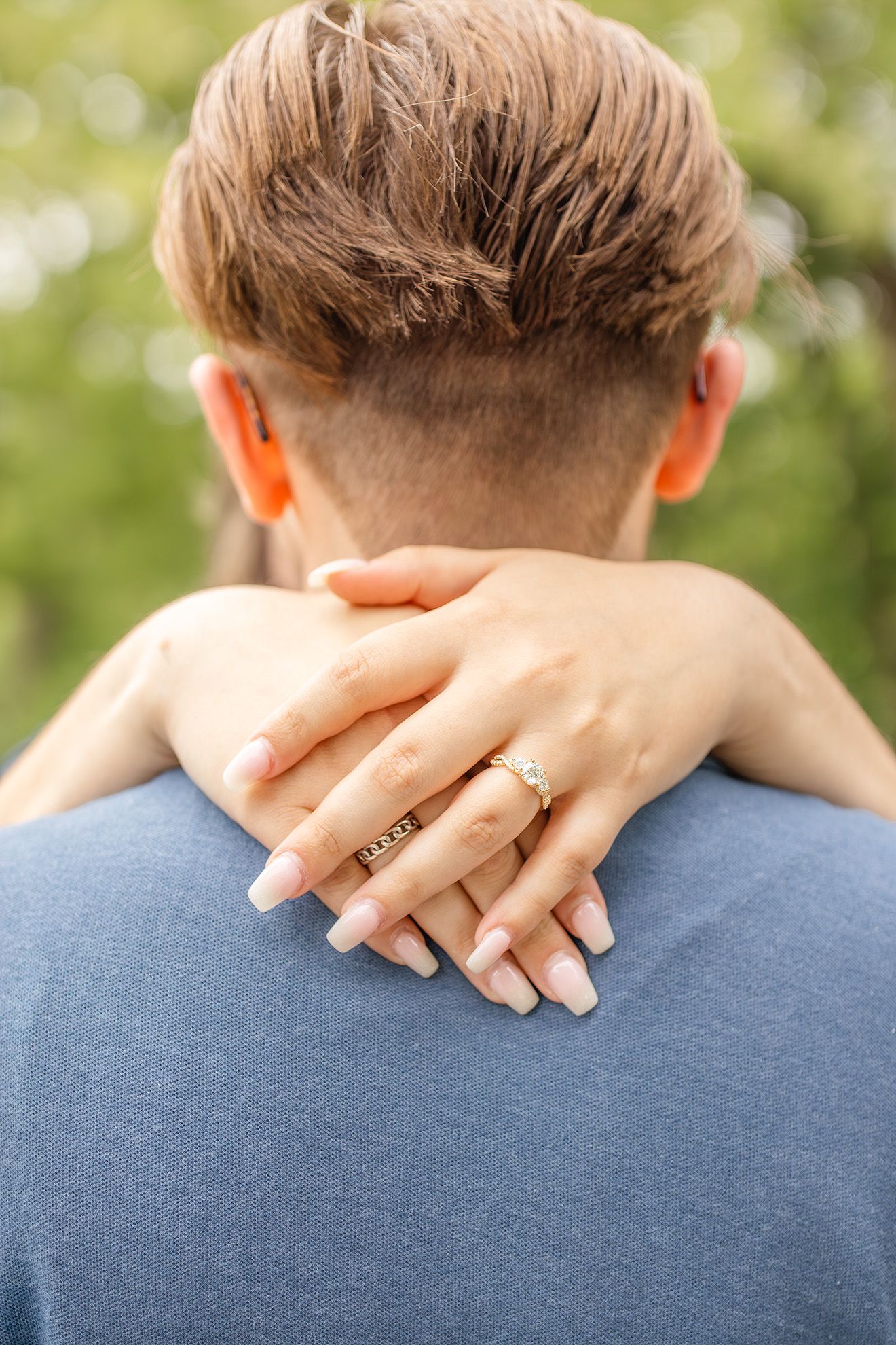 Close-up of a woman's hand with a gold ring resting on a man's shoulder from behind, outdoors.