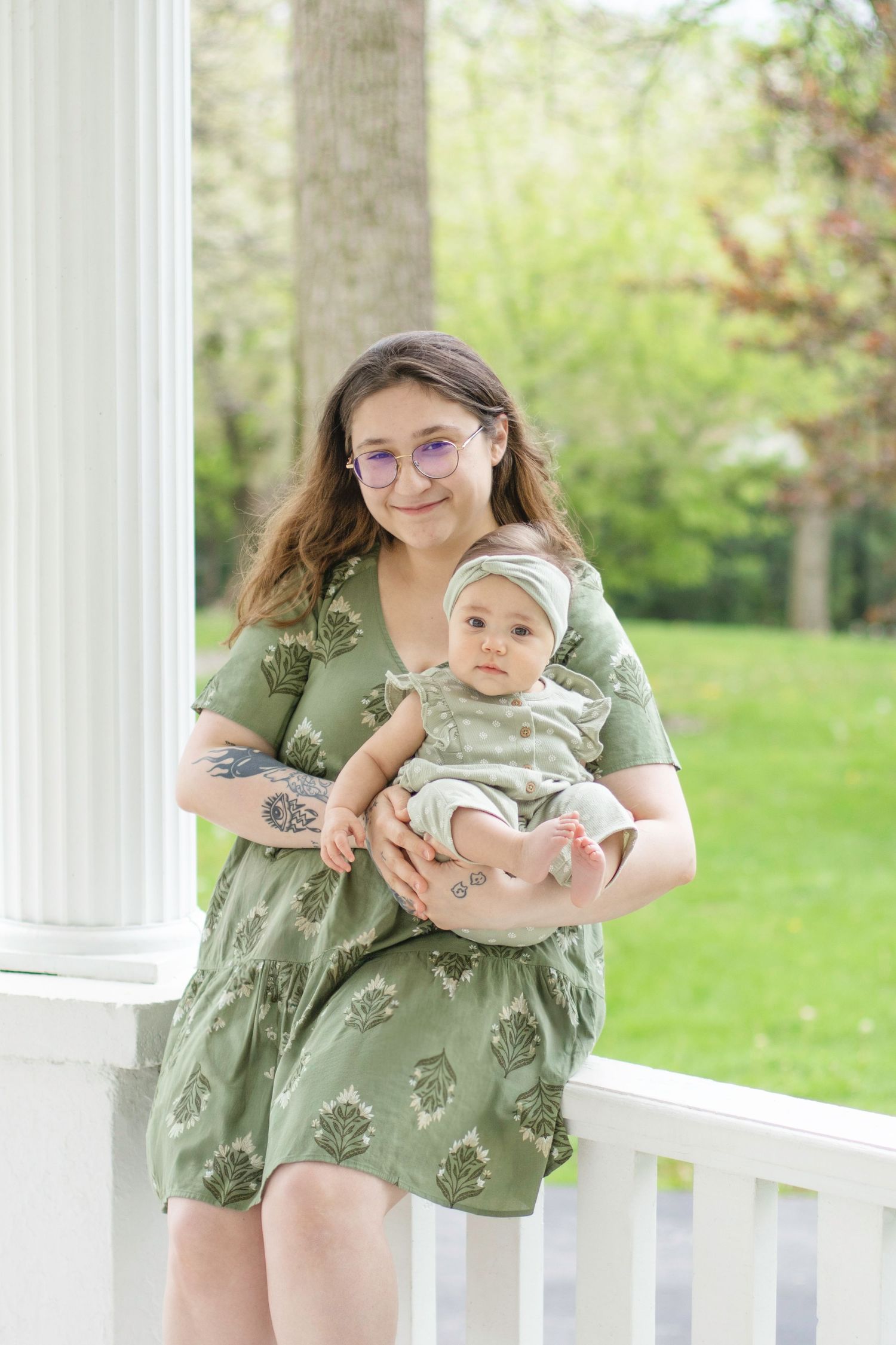 Mother and infant wearing coordinating sage green outfits smile together on white porch.