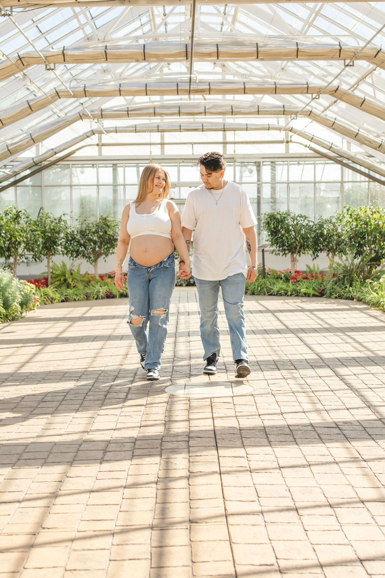 A couple walks hand in hand through a sunlit greenhouse filled with plants and flowers.