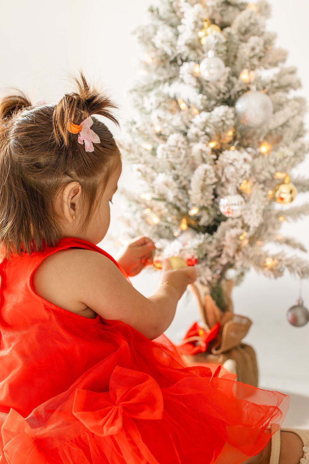 A child in a bright red tulle dress reaches up to decorate a snow-covered Christmas tree with twinkling lights.