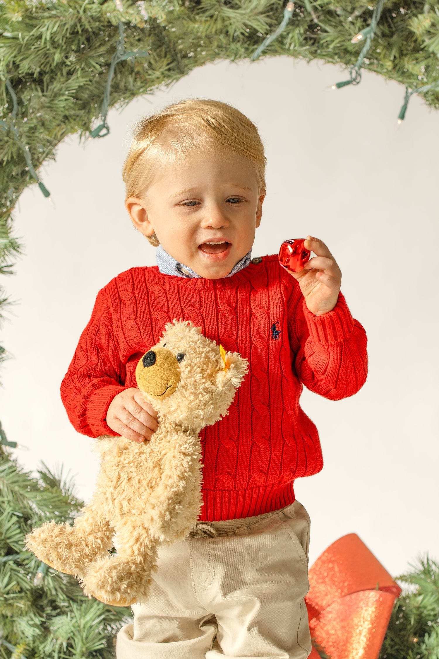 Small child in red cable knit sweater and khaki pants plays with plush teddy bear near holiday decorations.