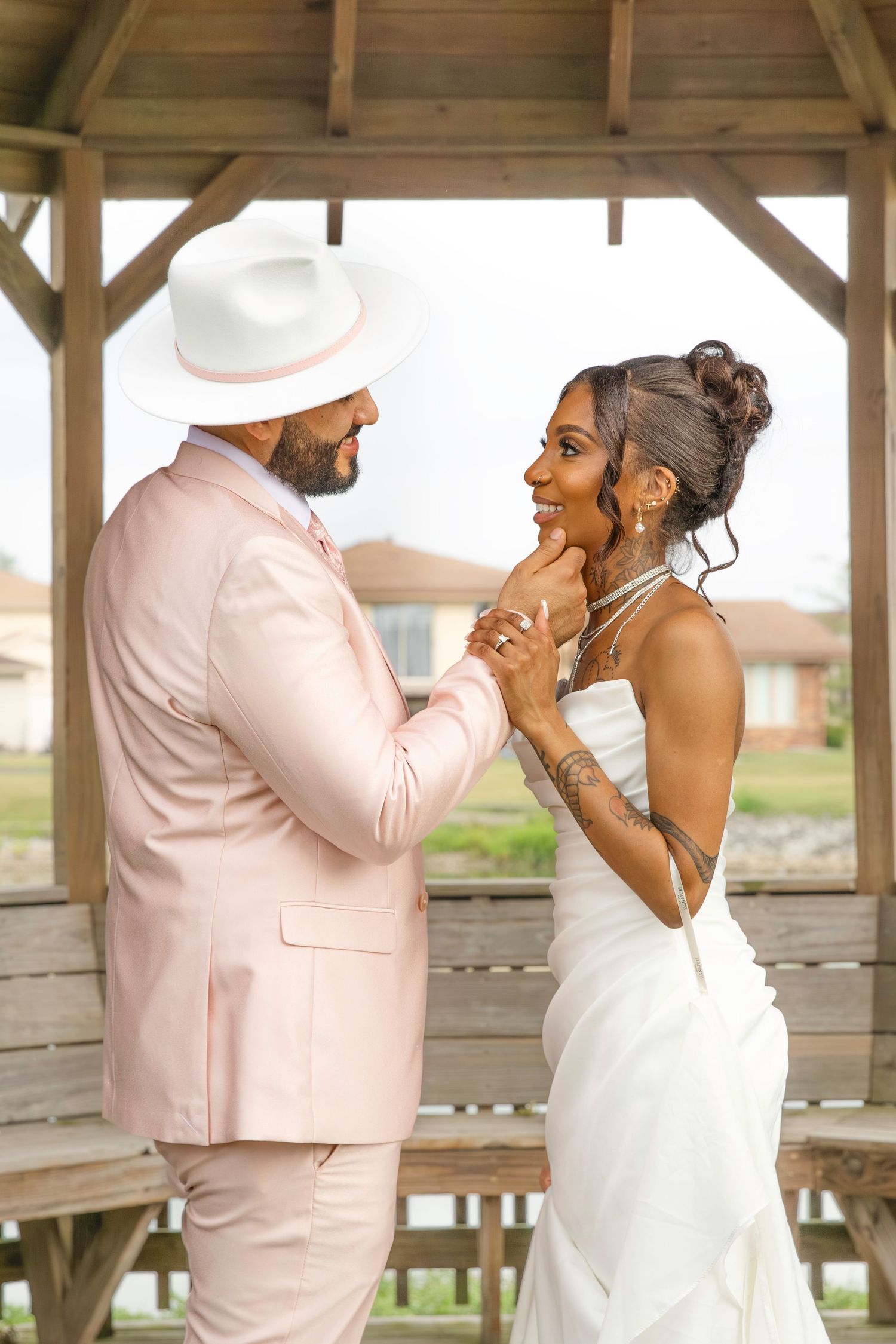 A couple in wedding attire, groom in pink suit and white hat, bride in white dress, share a tender moment under a wooden gazebo.