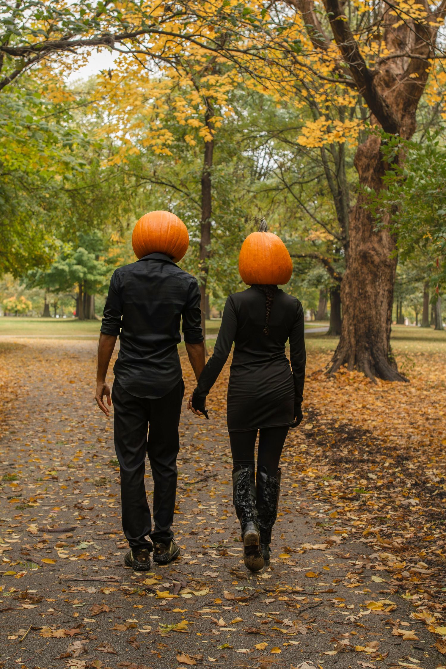 A couple wearing black outfits walks down an autumn path with pumpkins replacing their heads in a creative photo series.