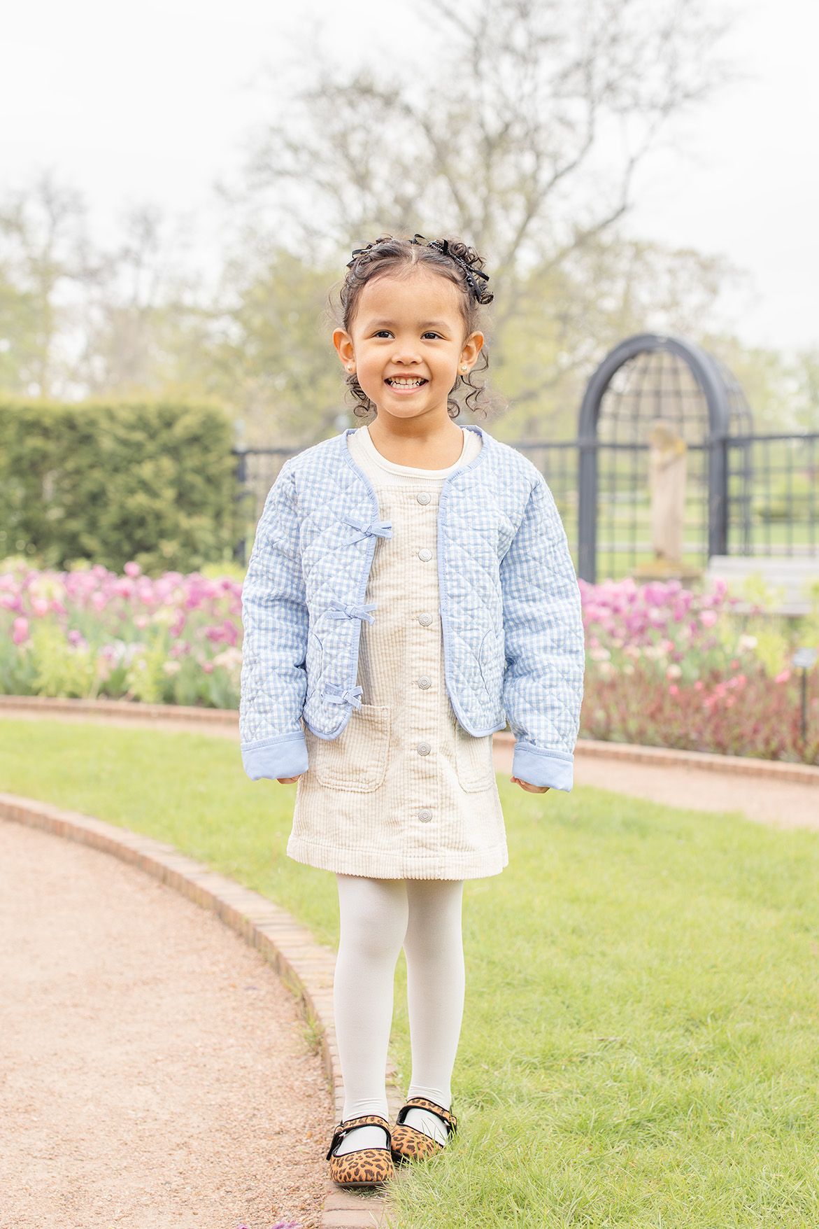 Smiling young girl in blue tweed jacket and cream dress standing on a garden path with colorful tulips behind her.