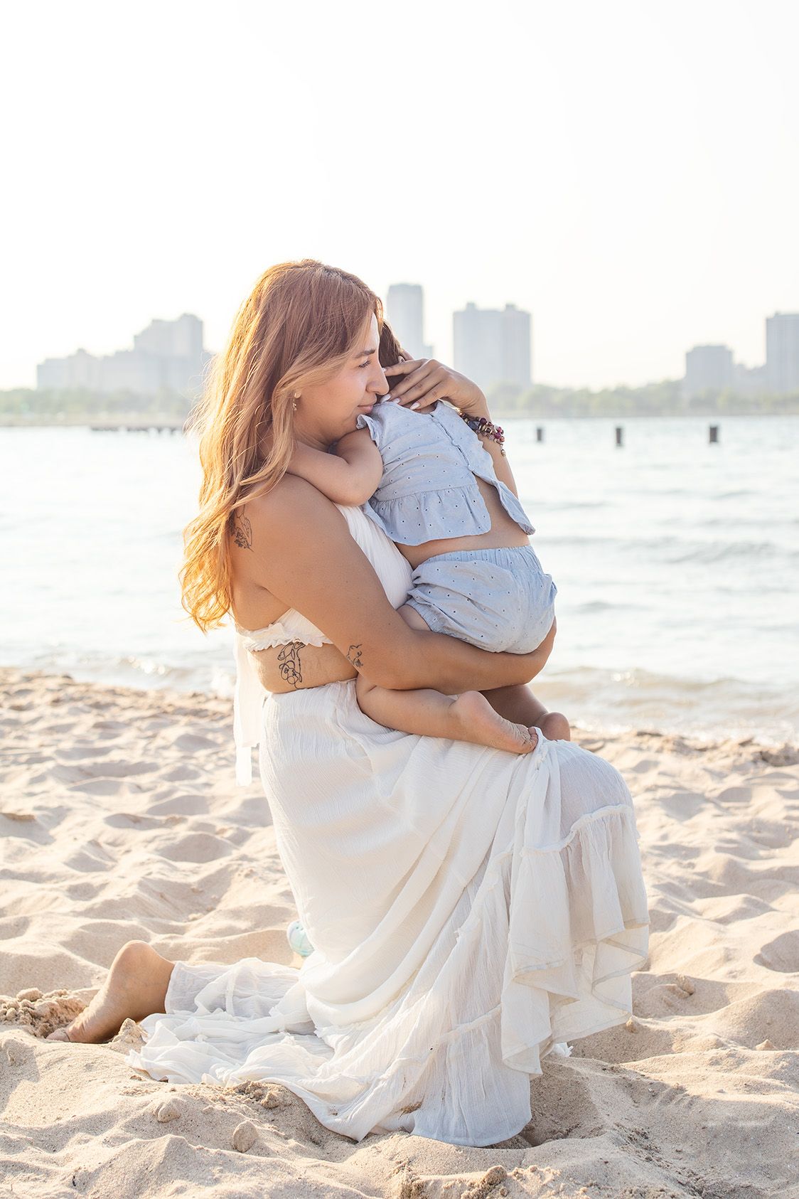 A woman in a white dress sits on a sandy beach, lovingly holding a baby, with a city skyline in the background.
